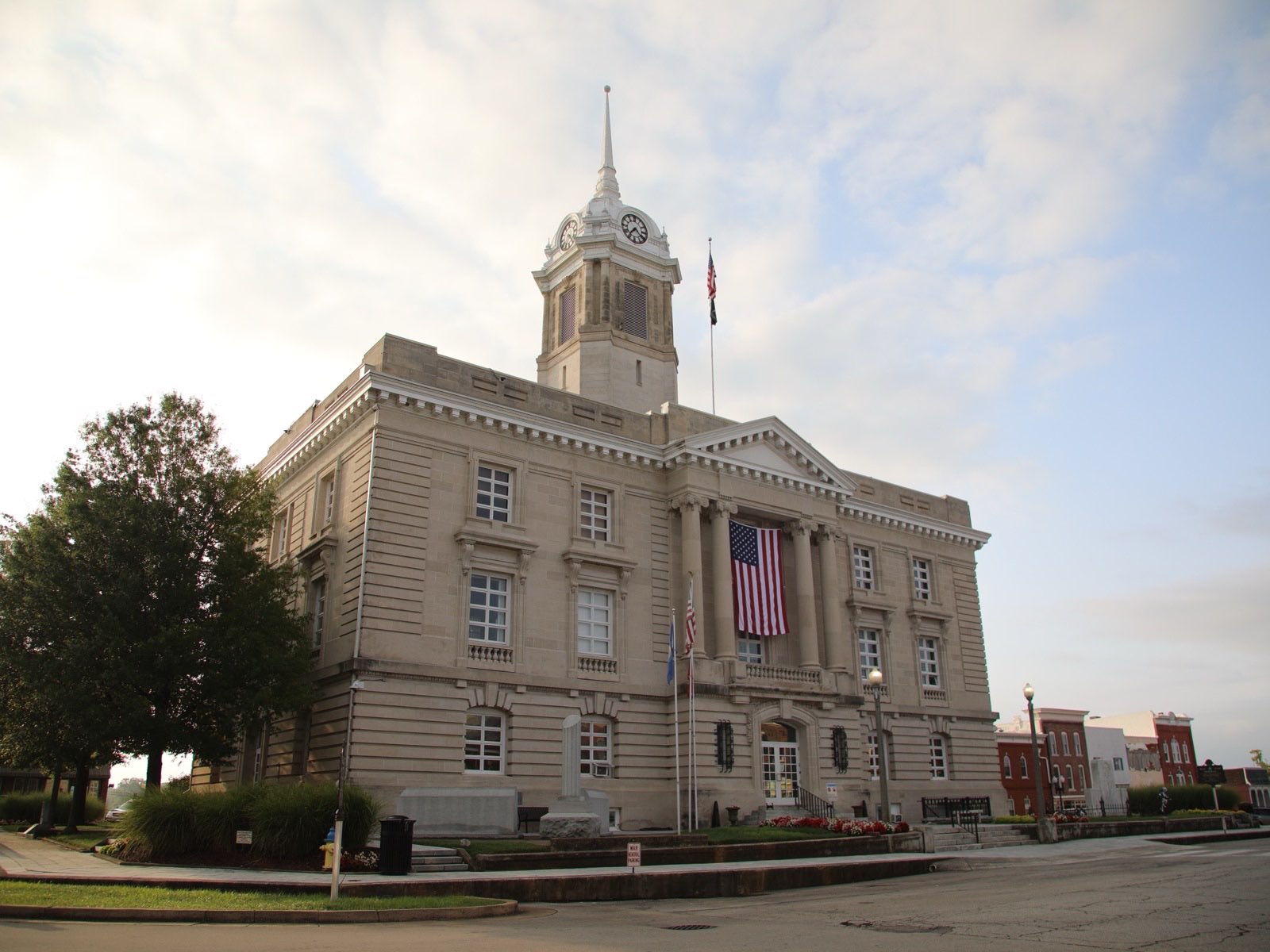 A three-storey limestone courthouse with a columned portico, a large American flag hung between the columns, and a white clock tower and spire above it, seen from the street corner