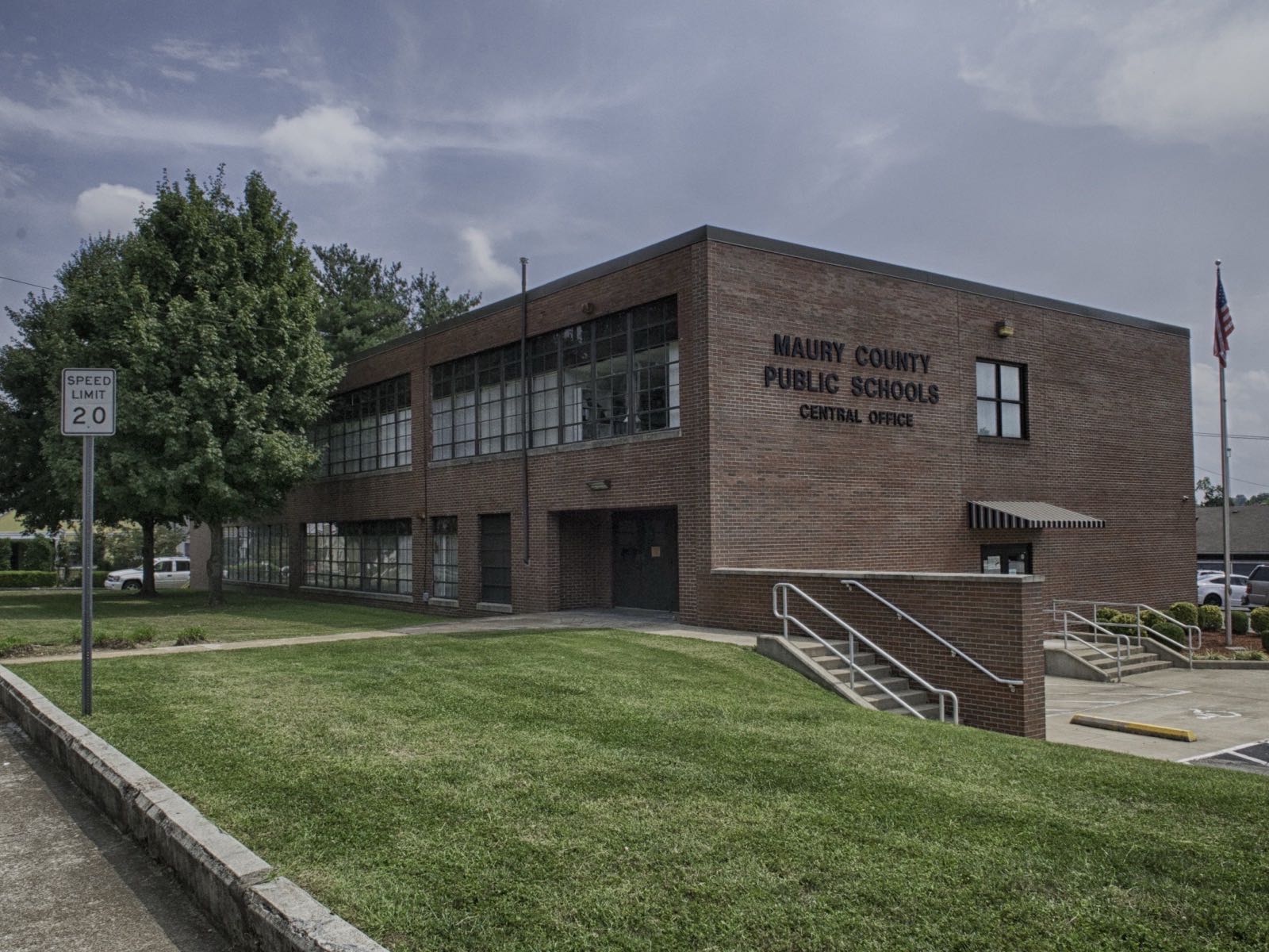 A two-storey mid-century red-brick building with a band of windows and lettering reading Maury County Public Schools Central Office, behind a mown lawn and a 20 mile-per-hour speed sign