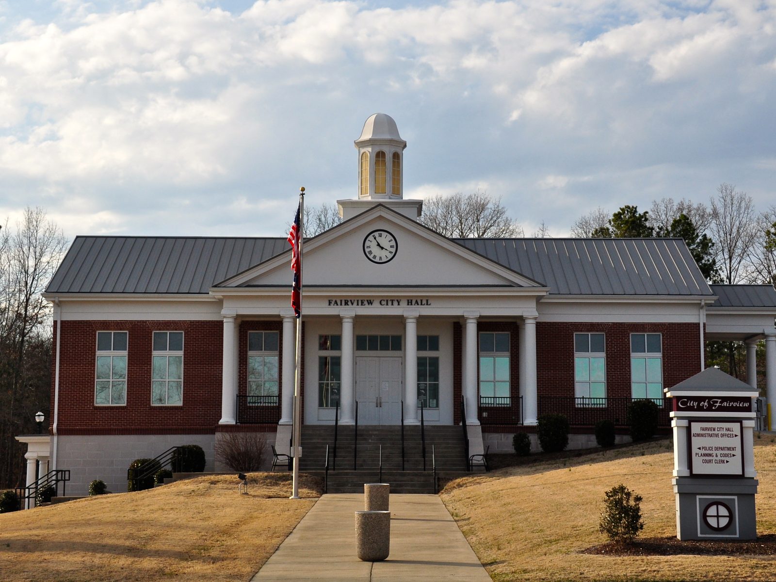 A one-story red-brick city hall with a white portico, a clock in the pediment, a cupola, and a flagpole on a dormant winter lawn under a cloudy sky