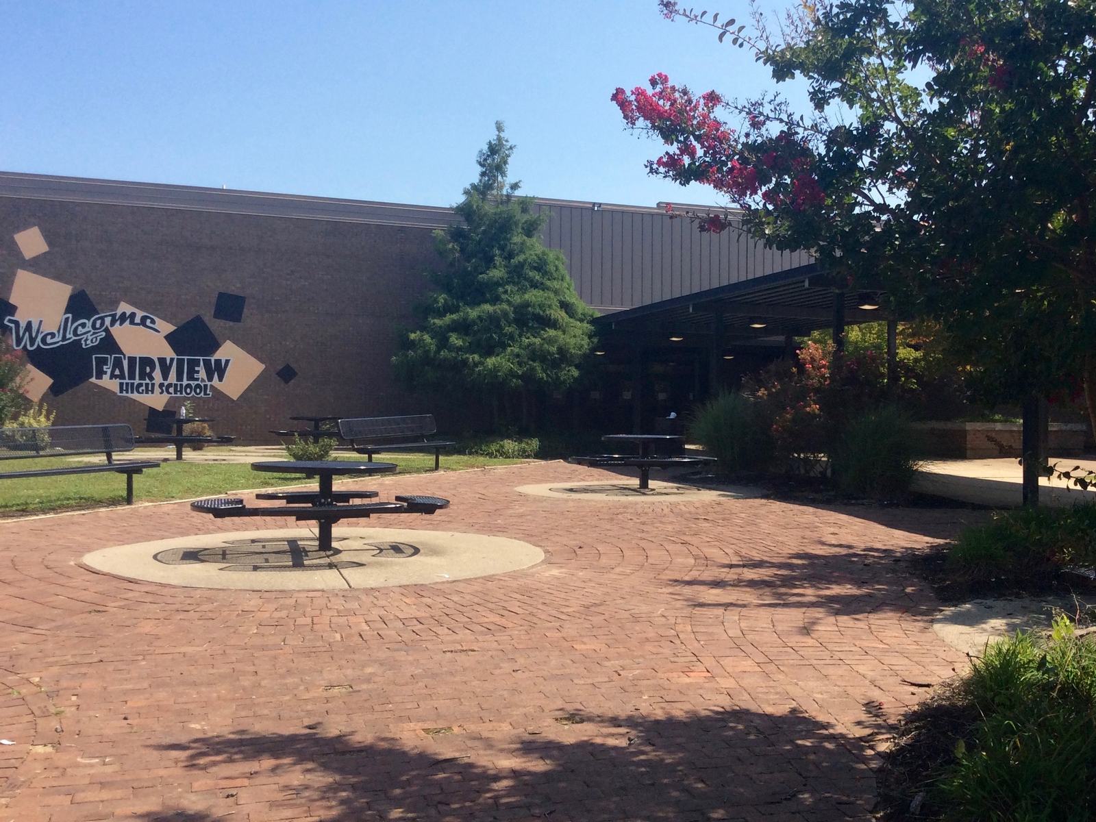 A brick-paved patio with round metal tables and benches in front of a tan brick school wall painted with a welcome to Fairview High School mural, a crepe myrtle in bloom