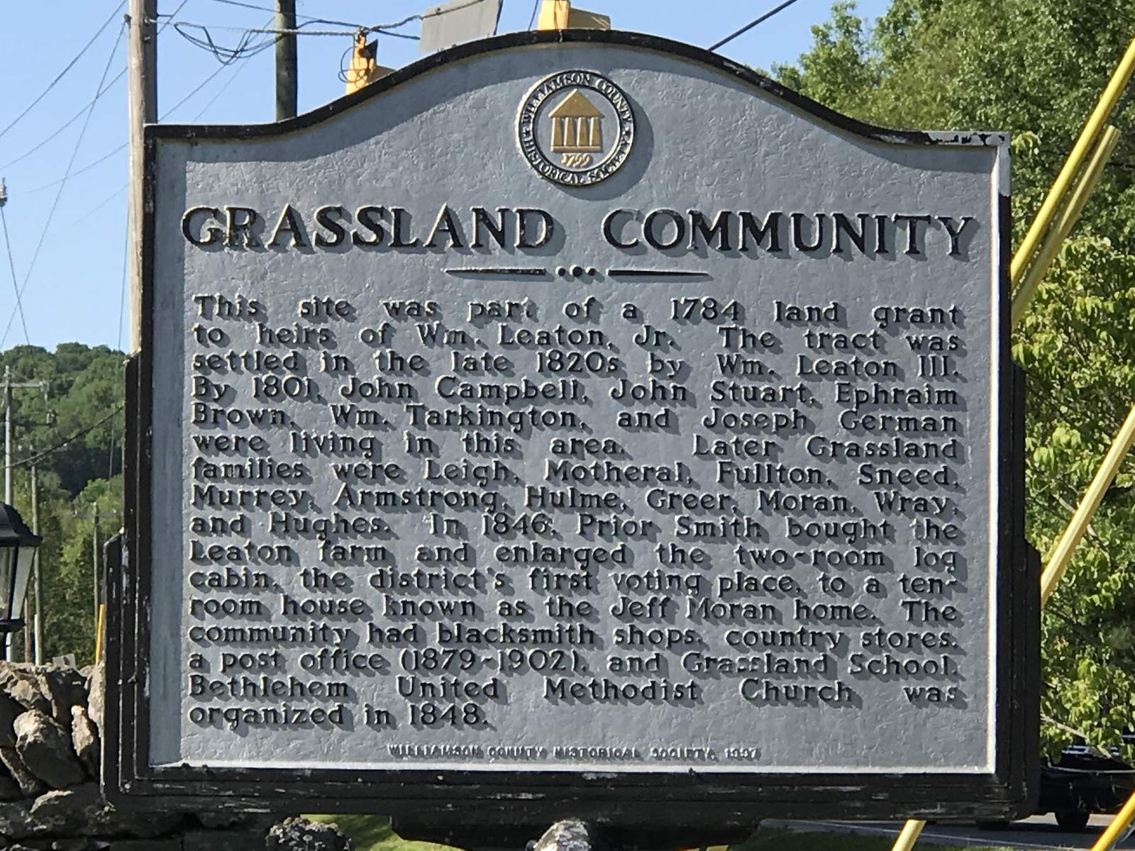 A cast-metal historical marker headed Grassland Community on a stone base beside a traffic signal, with trees and utility lines behind it