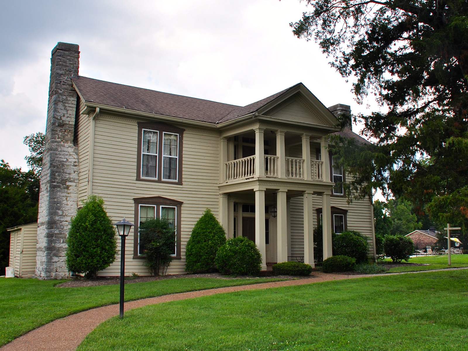 A two-story clapboard house with a stone end chimney, a two-tier columned porch, and clipped shrubs on a lawn under a large cedar