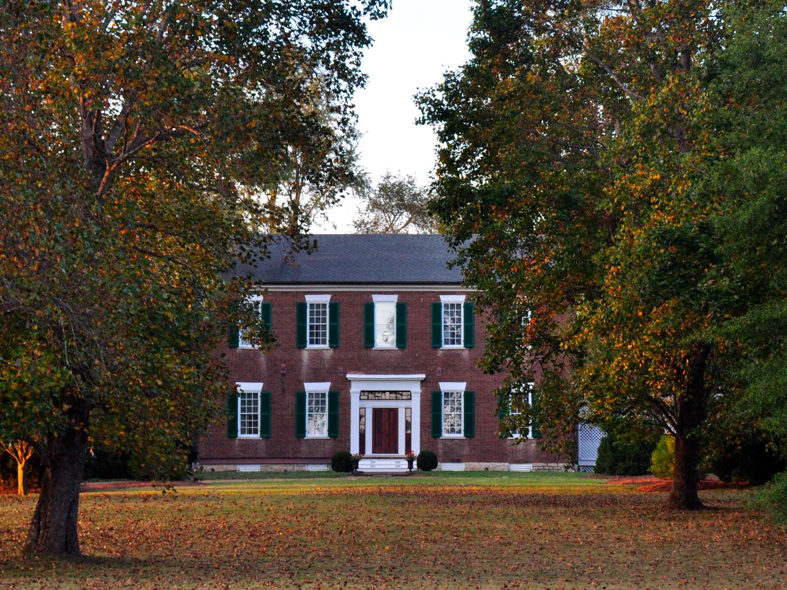 A two-story Federal brick house with green shutters and a white portico seen across a leaf-covered lawn between tall trees in autumn