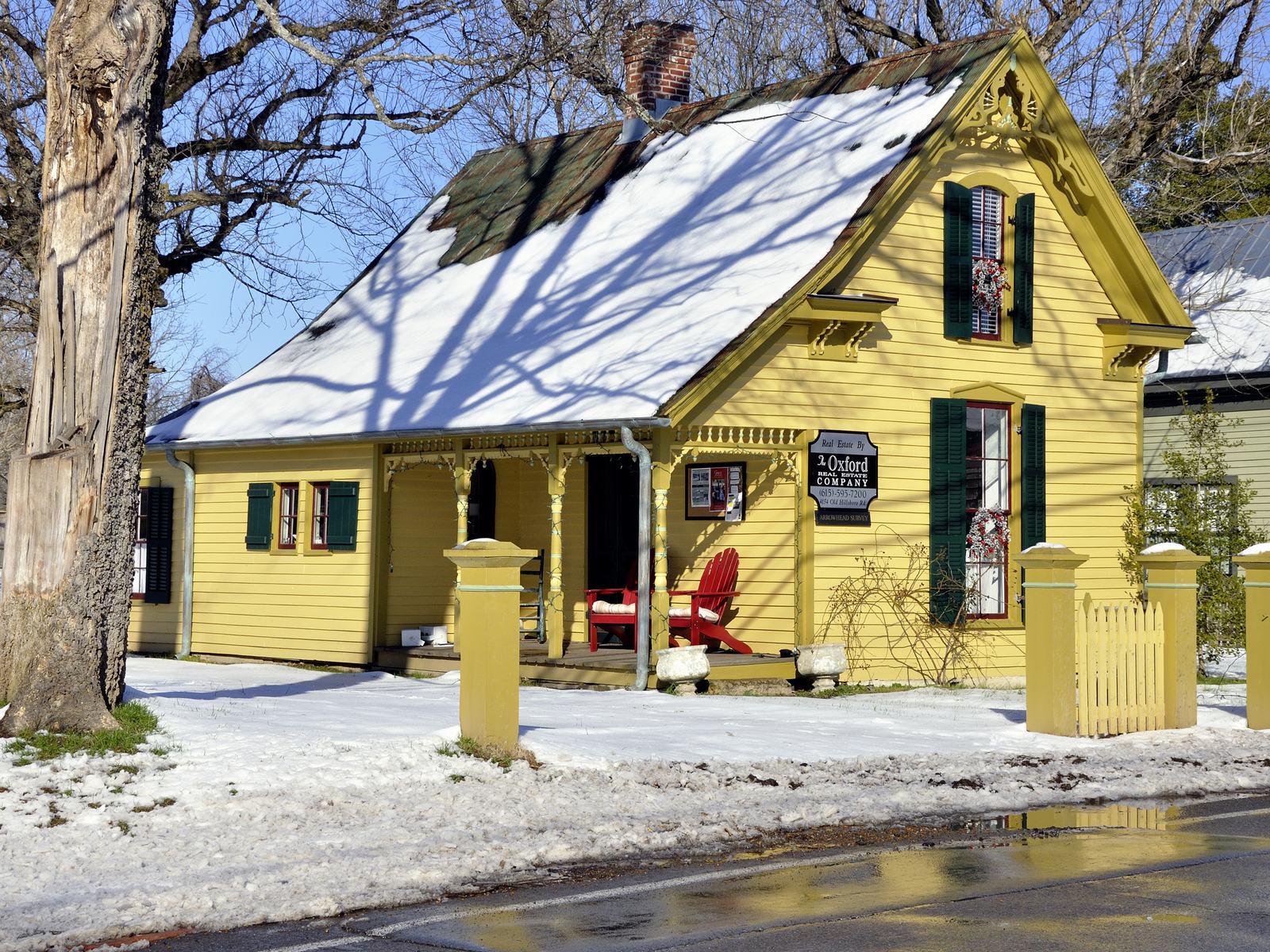 A yellow Gothic Revival cottage with green shutters, gingerbread trim, and a snow-covered tin roof behind a yellow picket fence on Old Hillsboro Road