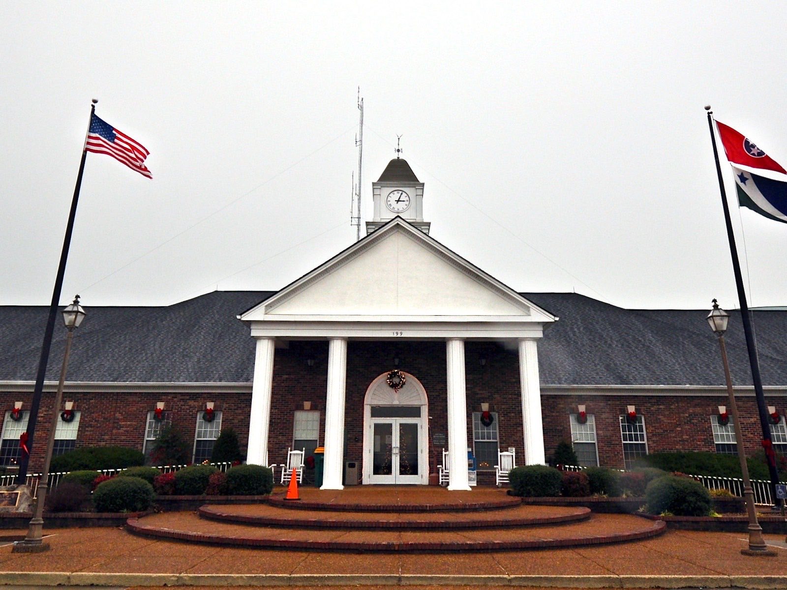 A long red-brick municipal building with a white columned portico, a clock cupola, and American and Tennessee flags on poles in front, decorated with Christmas wreaths