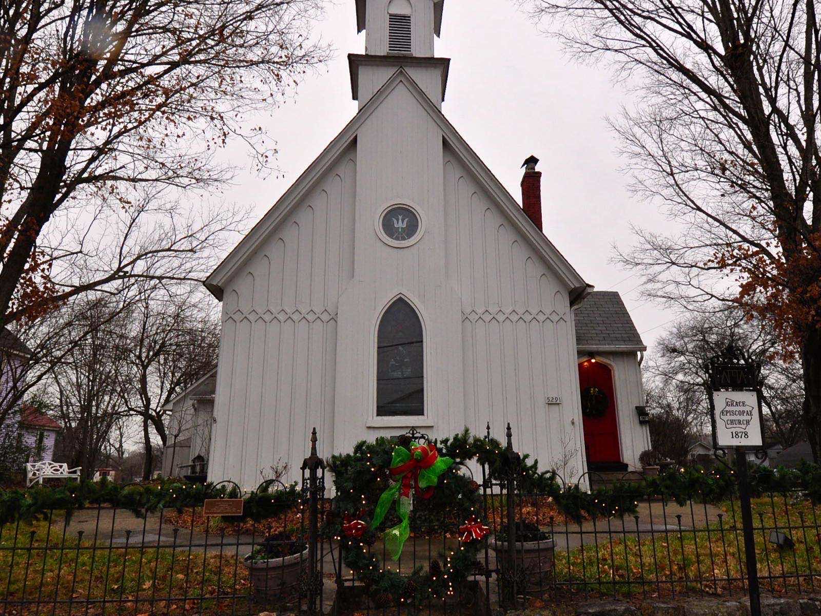 A small white board-and-batten Gothic Revival church with a steeple, red door and a stained-glass window, behind an iron fence hung with a Christmas wreath