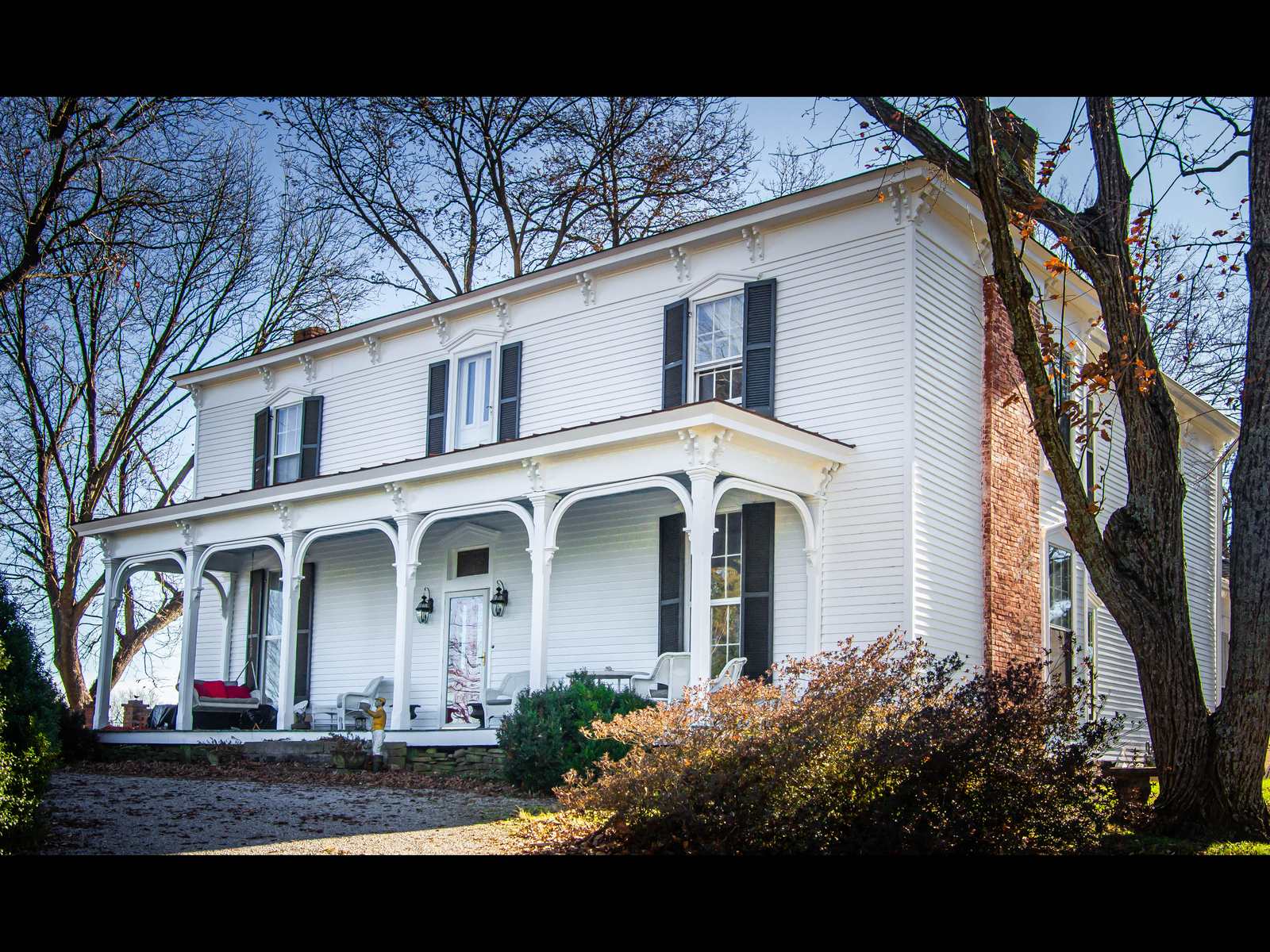 A two-story white frame farmhouse with a deep front porch and a metal roof, set behind a wooden fence and mature trees on a winter day